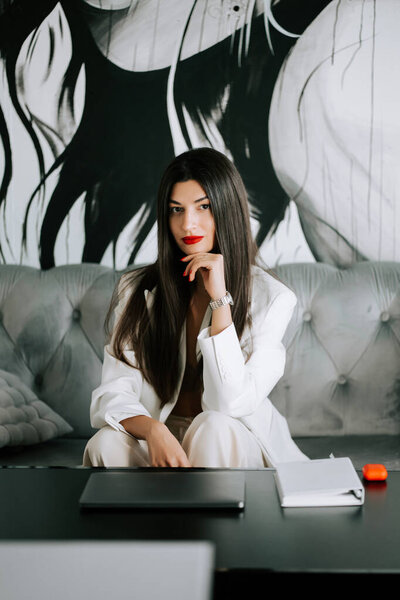 Young woman in white suit poses confidently in modern office setting with artistic background