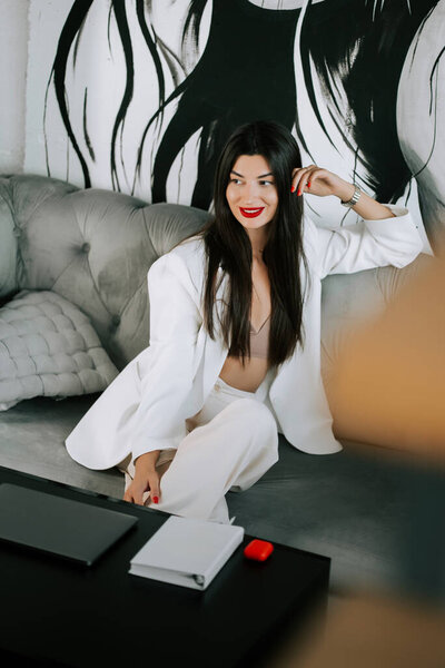 Elegant young woman in white outfit sitting on couch in modern space with artistic backdrop