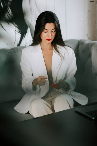 Woman in white suit sitting on couch engaging with a small object in a modern setting