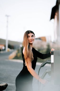 Kadın şarj istasyonunda gülümserken bir yandan da elektrik şarj aletini sıradanlaştırıyor.