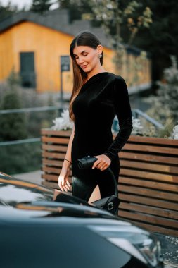 Siyah elbiseli zarif bir kadın, altın saat boyunca dışarıda lüks bir arabanın yanında poz veriyor.