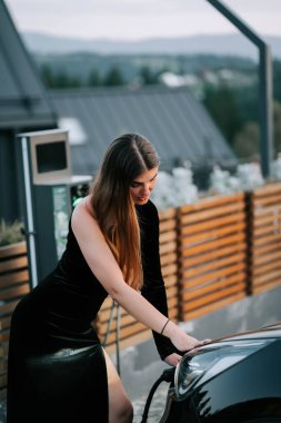 Zarif siyah elbiseli bir kadın açık hava manzarasında elektrikli araç yüklüyor.