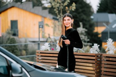 Gündüz vakti modern şehir ortamında genç bir kadın elektrikli araç şarj ediyor.