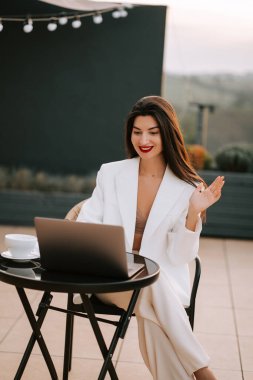 Şık beyaz elbiseli bir kadın gün batımında dışarıda bir kafede dizüstü bilgisayarla çalışıyor.