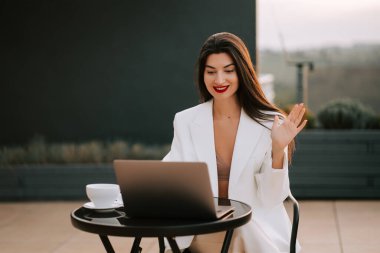 Güneşli bir öğleden sonra dışarıda görüntülü sohbet ederken gülümseyen ve el sallayan bir kadın.