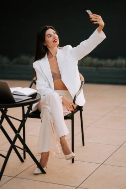 Genç bir kadın öğleden sonra laptopuyla açık kafede otururken selfie çekiyor.