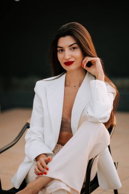 Modern bir ortamda zarif bir şekilde oturan kendine güvenen beyaz elbiseli bir kadın.