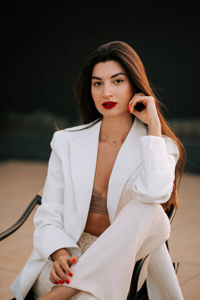 Woman in white blazer poses confidently with red lips in stylish outdoor setting at dusk
