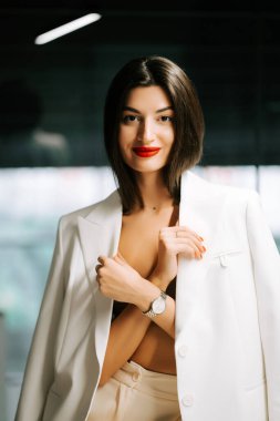 Beyaz ceketli zarif kadın modern ortamda güvenli bir şekilde içeride poz veriyor.