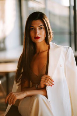 Zarif giyinmiş güzel bir kadın altın saatinde modern bir kafede güven içinde poz veriyor.