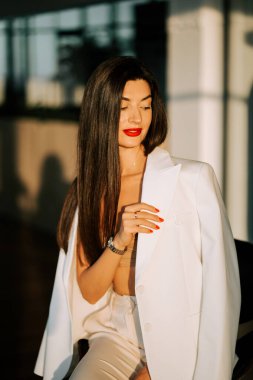 Beyaz ceketli şık bir kadın, altın saat boyunca kendine güvenen şık iç mekanlarda poz verir.