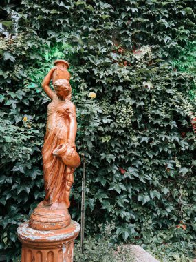 Bahçedeki yemyeşil sarmaşık zeminine çömlek taşıyan bir kadın heykeli.
