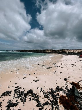 Kum kumsalı bulutlu gökyüzünün altında yumuşak dalgalar kıyıdaki karanlık kayalara çarpıyor.