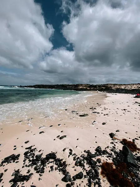 Kum kumsalı bulutlu gökyüzünün altında yumuşak dalgalar kıyıdaki karanlık kayalara çarpıyor.