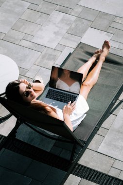 Genç bir kadın güneşli bir günde havuz kenarında dinlenirken dizüstü bilgisayarda çalışıyor.