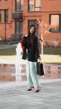 Modern bir parkta fıskiyelerin yanında yürüyen şık kıyafetler giyen ve alışveriş torbaları taşıyan bir kız.