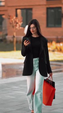 Modern bir şehir ortamında şık genç bir kadın alışveriş çantası ve telefonla dolaşıyor.