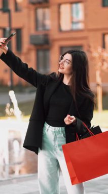 Genç bir kadın alışveriş yapmayı ve şehir meydanında parlak kırmızı çantalı selfie çekmeyi seviyor.