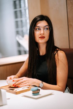 Modern bir kafe ortamında çay içen, şık dekorlu ve doğal ışıklandırmalı bir kadın.