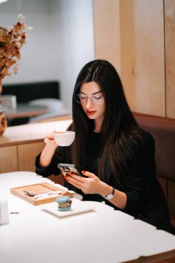 Genç bir kadın, rahat bir kafede akıllı telefon kullanırken kahveden zevk alıyor.