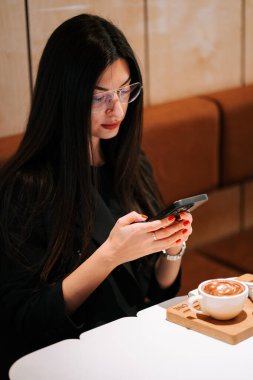 Genç bir kadın akıllı telefon kullanırken kahveden hoşlanır. 