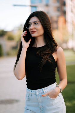 Young woman chatting on phone while walking in a sunny urban park during afternoon