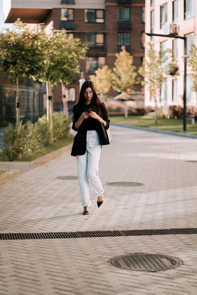 Young woman walks through urban area while texting on her phone during sunny afternoon