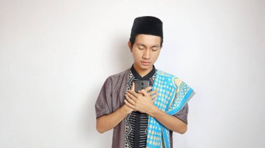 Asyalı Müslüman bir adam elini ve akıllı telefonunu göğsüne koyup beyaz bir arka plana koyuyor.