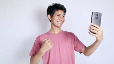 Akıllı telefonlu heyecanlı ve gülümseyen Asyalı adam izole beyaz arka planda el kol hareketi yapıyor.