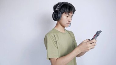 ciddi Asyalı adam kulaklık kullanıyor ve beyaz arka planda cep telefonu oyunları oynuyor.