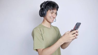 heyecanlı Asyalı adam kulaklık kullanıyor ve beyaz arka planda akıllı telefon çalıyor.