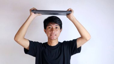 Genç Asyalı mutlu bir jest yapıyor. Kafasının üstünde beyaz bir arka planda dizüstü bilgisayar taşıyor.