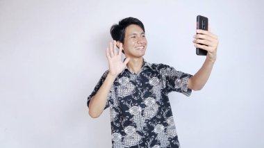 Batik giyen yakışıklı genç Asyalı bir adam görüntülü arama yapıyor, izole edilmiş beyaz bir arka planda akıllı telefon kullanarak selfie çekiyor.