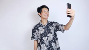 Batik giyen yakışıklı genç Asyalı bir adam görüntülü arama yapıyor, izole edilmiş beyaz bir arka planda akıllı telefon kullanarak selfie çekiyor.