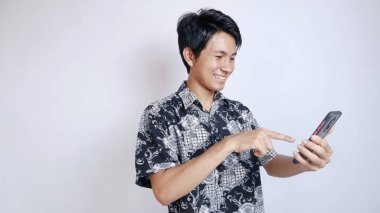 Batik giyen yakışıklı genç Asyalı bir adam görüntülü arama yapıyor, izole edilmiş beyaz bir arka planda akıllı telefon kullanarak selfie çekiyor.
