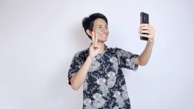 Batik giyen yakışıklı genç Asyalı bir adam görüntülü arama yapıyor, izole edilmiş beyaz bir arka planda akıllı telefon kullanarak selfie çekiyor.