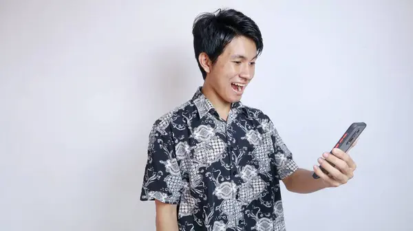 stock image Handsome young Asian man wearing batik is making a video call, taking a selfie using a smartphone on an isolated white background