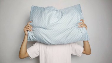 Asyalı genç adam yüzünün önünde bir yastıkla poz veriyor. Böylece tüm yüzünü izole edilmiş beyaz bir arka planda kapatıyor.