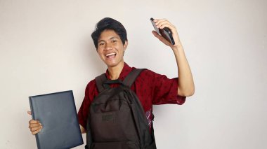 Batik tişörtlü yakışıklı Asyalı bir adam bir çanta taşıyor, elinde bir kitap ve akıllı telefon, neşe, mutluluk, coşku ifade ediyor, izole edilmiş beyaz bir arka planda zafer.