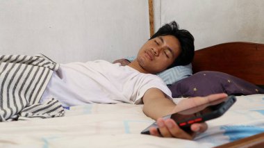 A young Asian man in a white shirt is asleep on the bed and still holding a smartphone clipart