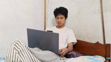 Genç Asyalı adam yatağın üstünde dizüstü bilgisayar ve akıllı telefon kullanıyor.