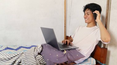 Genç Asyalı erkekler oynuyor, kullanıyor, izliyor, video görüşmeleri yapıyor, dizüstü bilgisayarla tartışıyor ve yatakta telefon görüşmesi yapıyorlar.