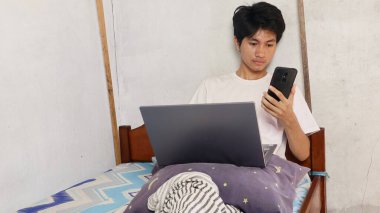 Genç Asyalı erkekler yatağın üzerinde dizüstü bilgisayar ve akıllı telefon kullanarak oynar, kullanır, izler, izler, video görüşmesi yapar, tartışır.