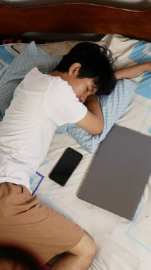 Genç Asyalı bir adam laptopuyla yatakta uyuyor.