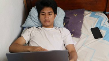 Genç Asyalı adam yatak odasında uzanmış dizüstü bilgisayarını izliyor ve oynuyor, beyaz bir gömlek giyiyor.