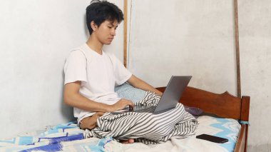 Beyaz tişörtlü genç Asyalı yatakta dizüstü bilgisayar kullanıyor, çalışıyor ya da bilgisayarda oynuyor.