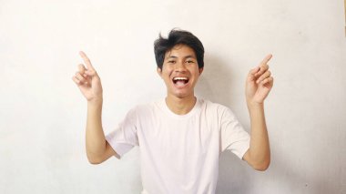 Genç Asyalı mutlu poz verirken beyaz bir arka planı işaret ediyor, beyaz gömlek giyiyor.