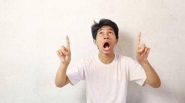Genç Asyalı adam beyaz gömlekli, beyaz bir arka planı işaret ederken şaşırmış görünüyor.