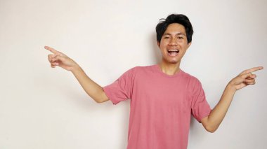 Heyecanlı genç yakışıklı Asyalı mutlu, neşeli ve coşkulu pozlar soldan ve sağdan izole edilmiş beyaz bir arkaplanı işaret ediyor. Kırmızı giysiler giyiyor.