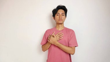 Genç Asyalı bir adam, beyaz bir arka planda göğsünü acı içinde tutarak poz veriyor. Kırmızı giysiler giyiyor.